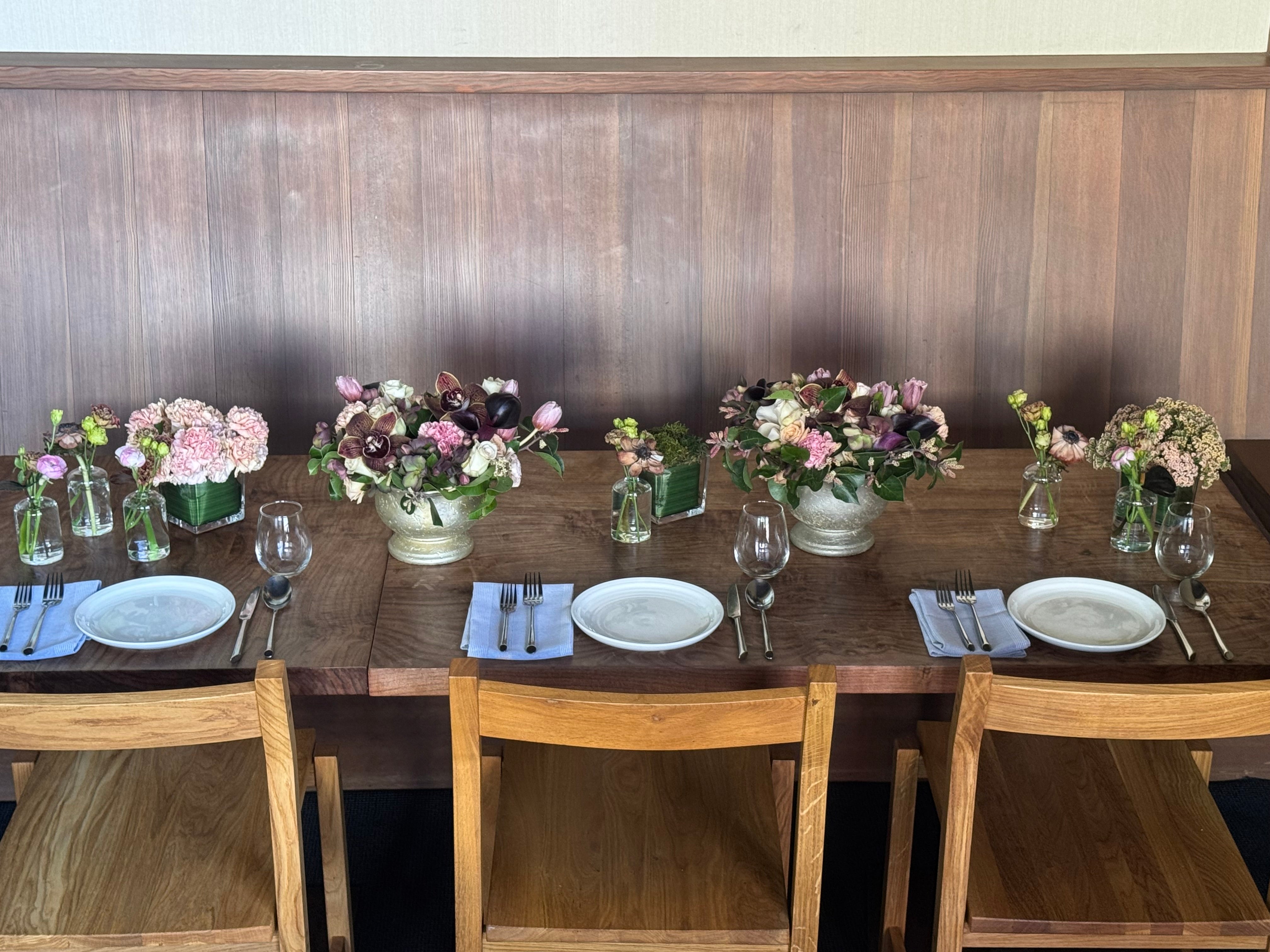 A rustic wooden dining table set for four, featuring white plates, silver cutlery, wine glasses, folded blue napkins, and small floral centerpieces in glass vases, set against a wood-paneled wall.