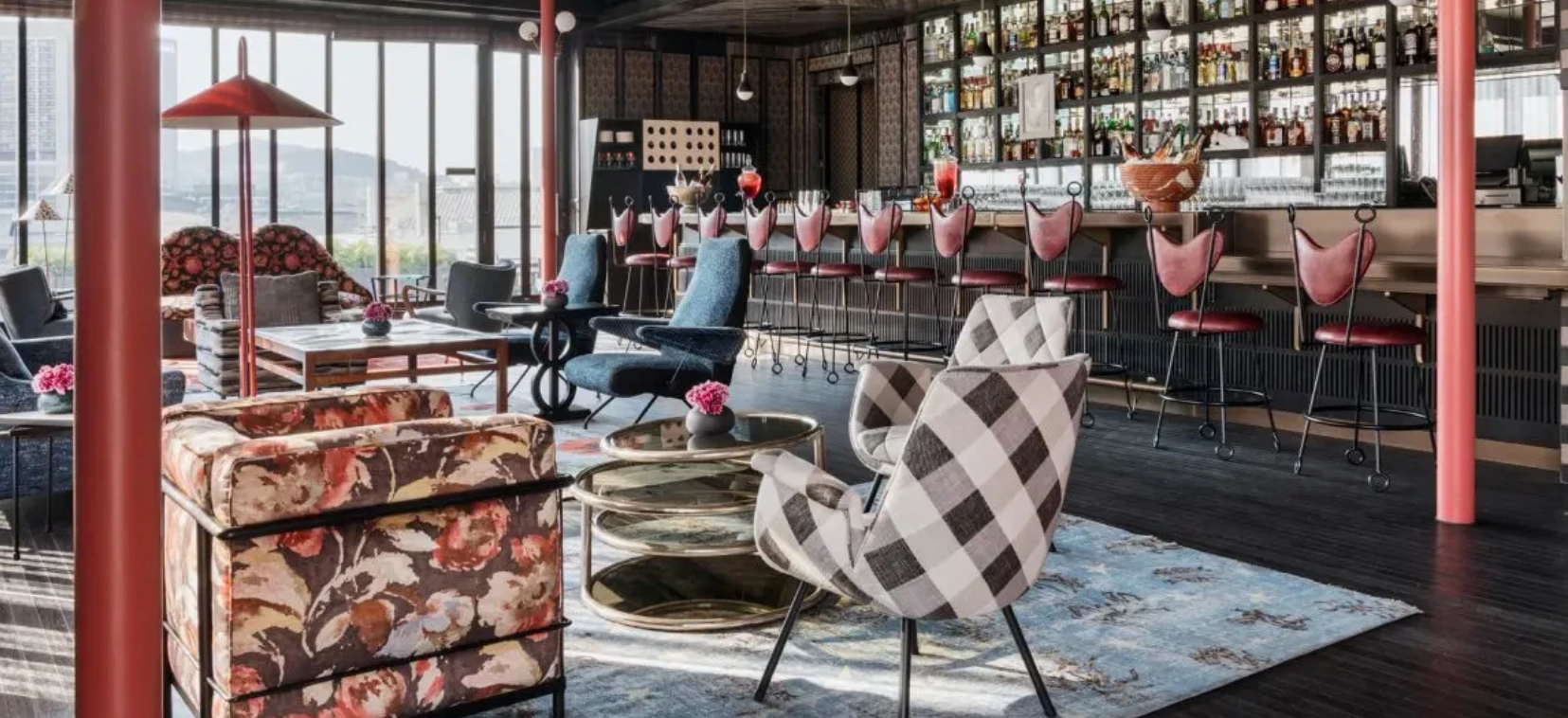 A stylishly decorated lounge bar interior with patterned armchairs, a floral sofa, a bar counter lined with pink bar stools, and a large back wall of liquor bottles. Large windows on the left offer a view of outdoor hills.