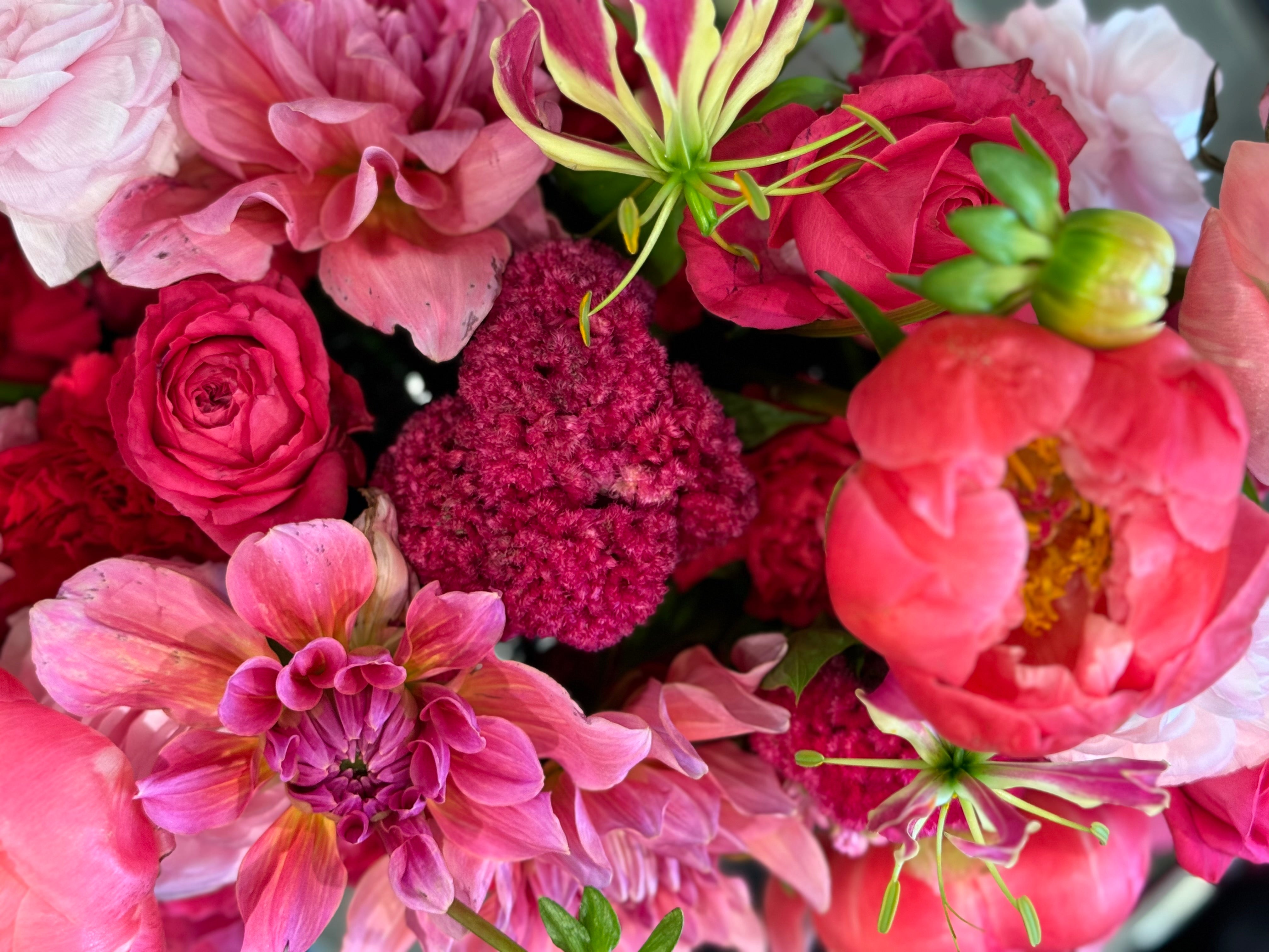 A close-up of a vibrant floral arrangement featuring pink and red roses, a pink peony, a magenta blooms like dahlias, roses, glories, sweet pea, orchids, pale pink carnation.