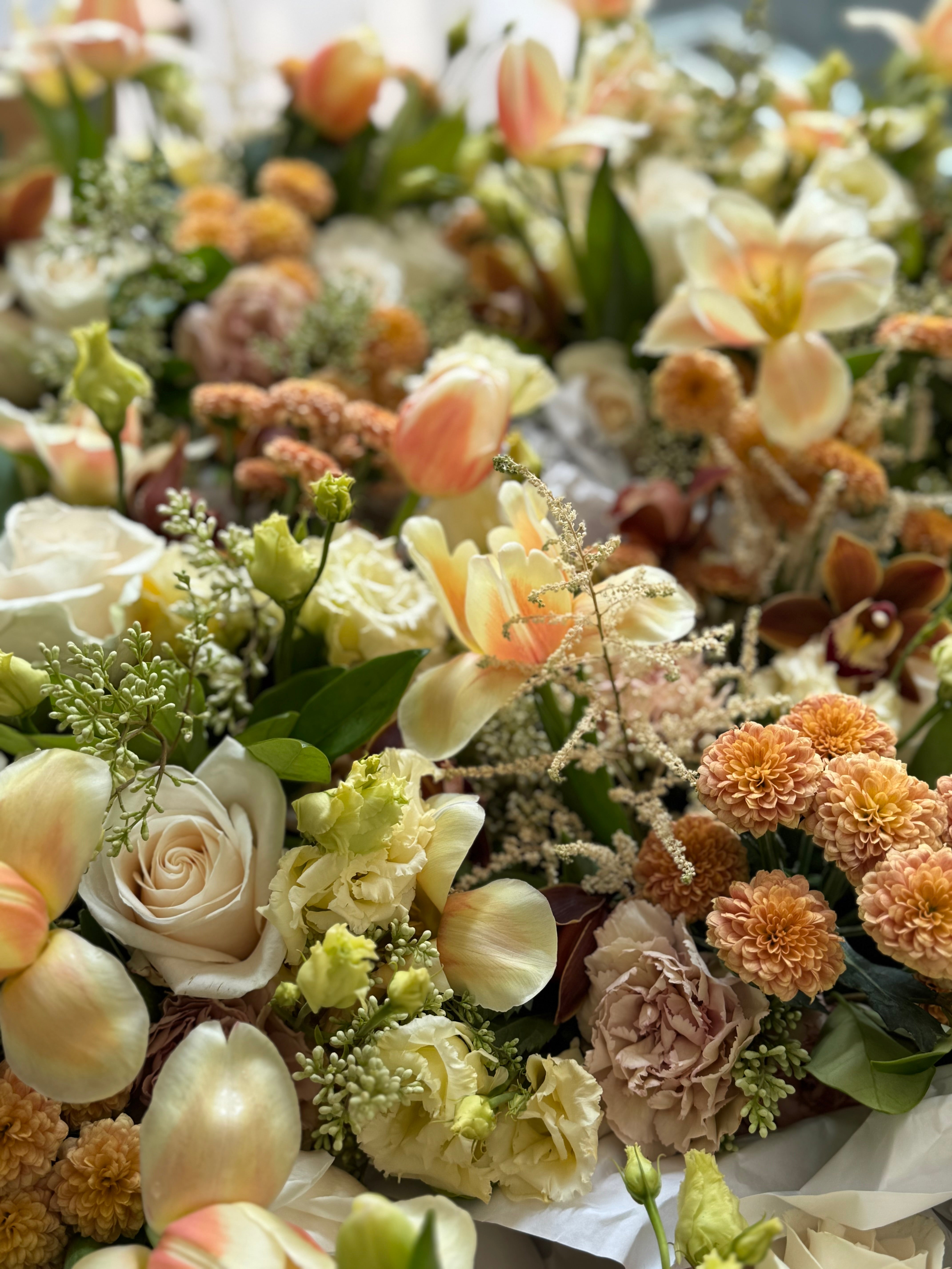 A close-up of a mixed floral arrangement featuring cream roses, peach tulips, orange chrysanthemums, and green foliage.