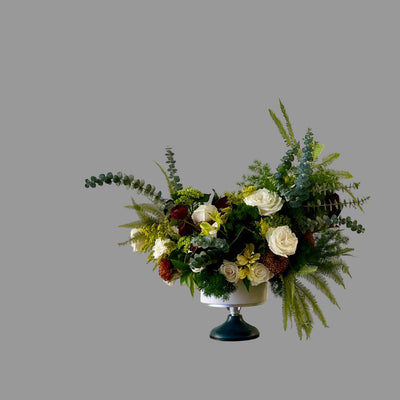 A floral arrangement in a white pedestal bowl with a dark teal base, set on a gray stone block against a plain light beige wall. The bouquet features white and cream roses, green eucalyptus and ferns, and small red and yellow flowers.