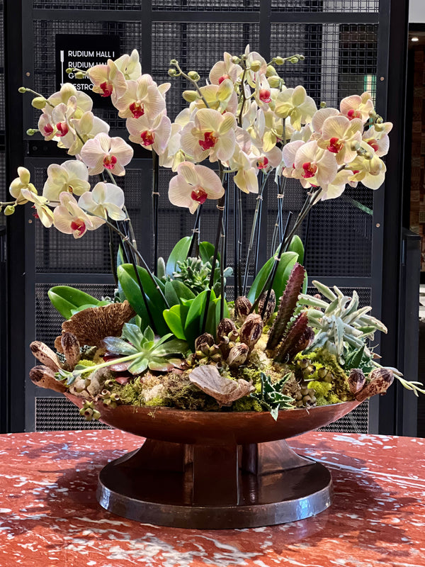 A large decorative arrangement of pale yellow and white orchids with red centers, green leaves, and assorted succulents and moss in a copper-toned bowl pedestal planter, displayed on a red marble tabletop in front of a black metal mesh wall.