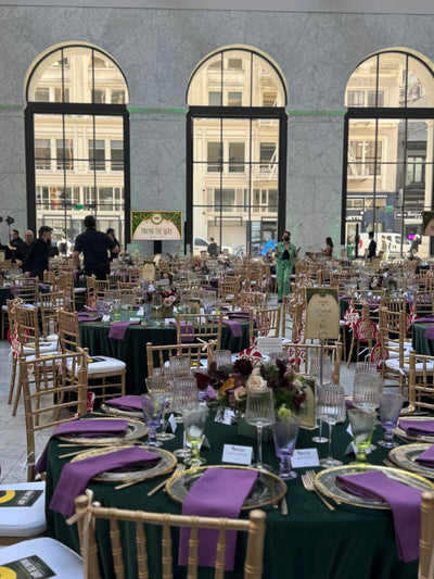 A large event hall with three tall arched windows, filled with round tables set for guests. The tables have dark green tablecloths, purple napkins, gold-rimmed plates, and floral centerpieces.