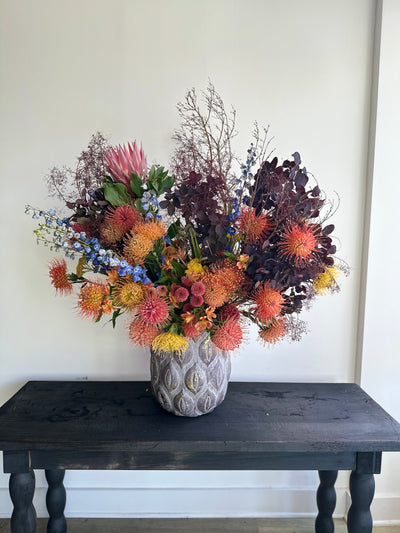huge swoop floral arrangement of protea and smoke bush in oranges, golds, yellows and dark purple in a huge concrete pot
