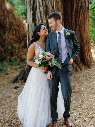 the couple poses in front a huge redwood tree and the bride holds and organic modern flower arrangement full of all sorts of nice things